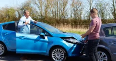 driver standing by teal-blue car facing driver of a dark blue car with rear-end damage