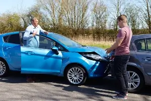 driver standing by teal-blue car facing driver of a dark blue car with rear-end damage